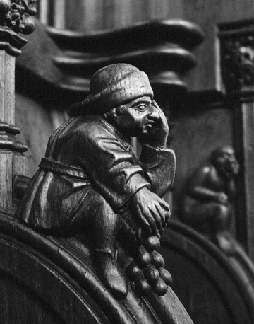 Koorbanken, detail: zittende man met omhoog getrokken knieen en een mand tussen zijn benen geklemd