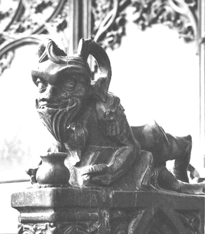 Koorgestoelte, detail: liggend figuurtje,  voorstelling van duivel met attributen van de bok: baard, hoorns en bokkepoten (vooraanzicht)