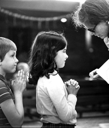 Twee kinderen met gevouwen handen, tegenover een vrouw (gebogen)